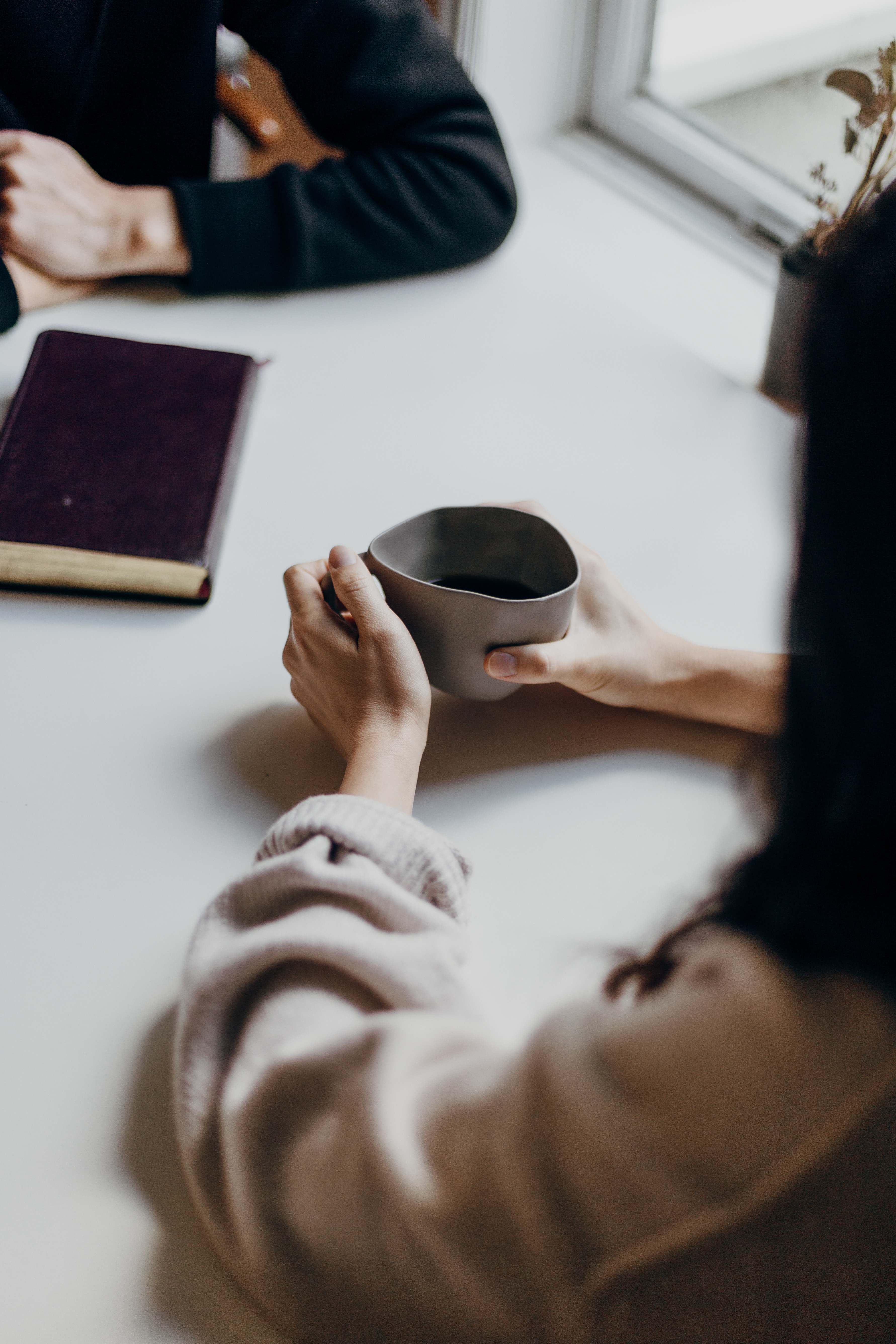 Photo of two people at a table talking. There is a book on the table and one person is holding a coffee cup.