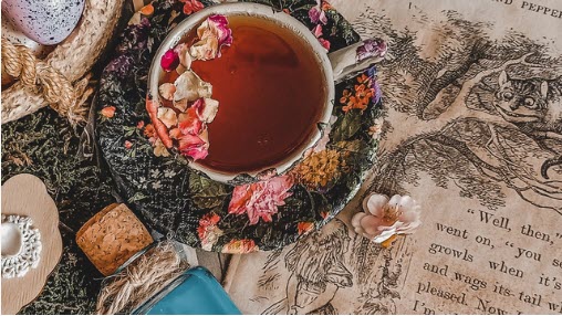 Tea in china cup and saucer resting on open ALice In Wonderland book, bottle on its side with blue liquid, dried flowers in tea and around the cup